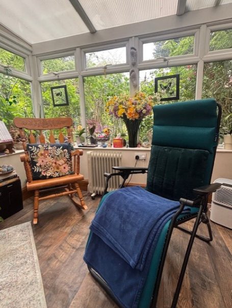 Mindful Moments Therapies A cosy conservatory featuring a green chair, a rocking chair, and a vase of flowers.