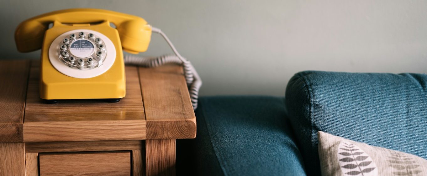 Contact Phiona A bright yellow rotary telephone on a wooden side table beside a blue sofa.