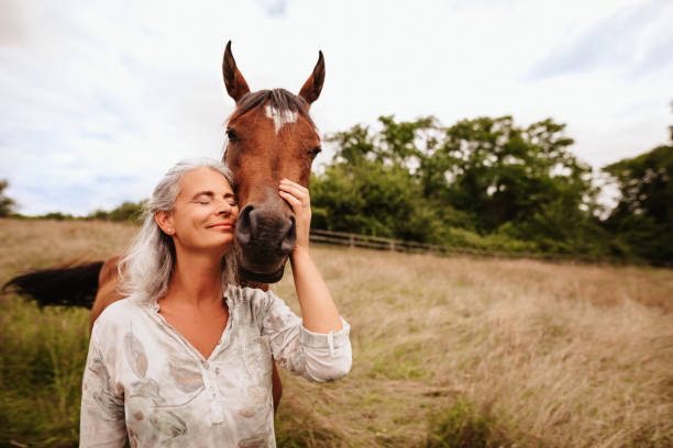 Equine Assisted Therapy Woman gently embraces a horse, both smiling in a grassy field under a cloudy sky.